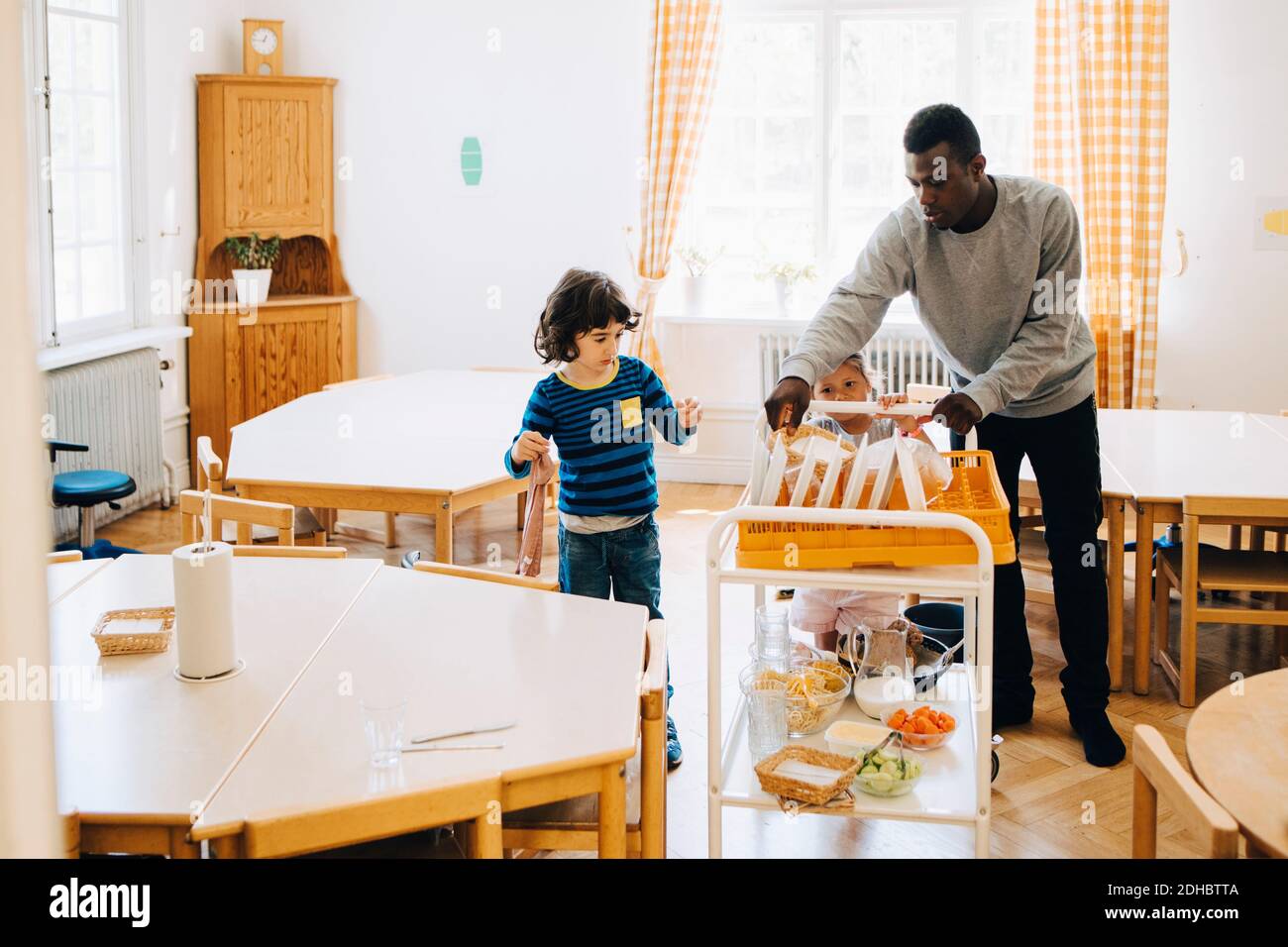 Male teacher assisting students pushing cart amidst tables in classroom at child care Stock Photo