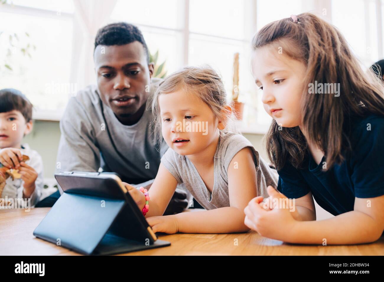 Male teacher looking at girl using digital tablet by friend at table in classroom Stock Photo