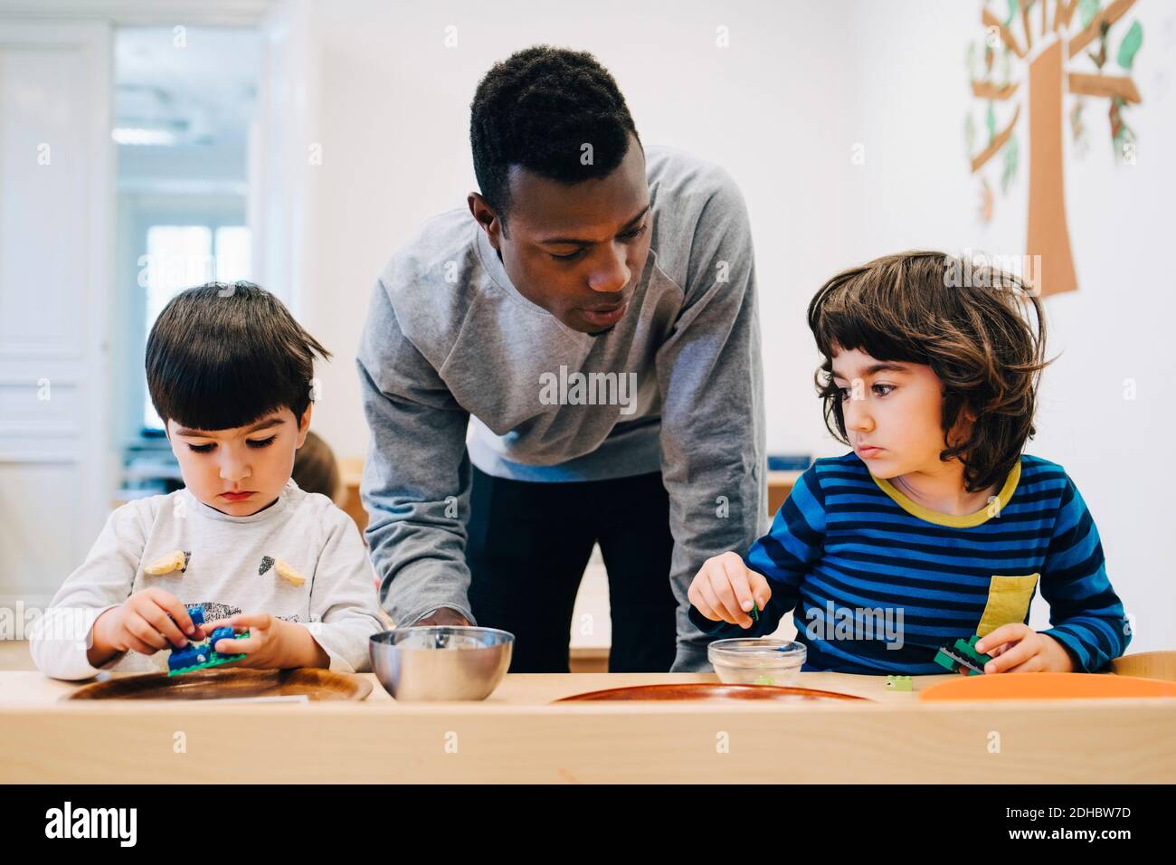 Male teacher looking at students playing with toys at table in child care classroom Stock Photo