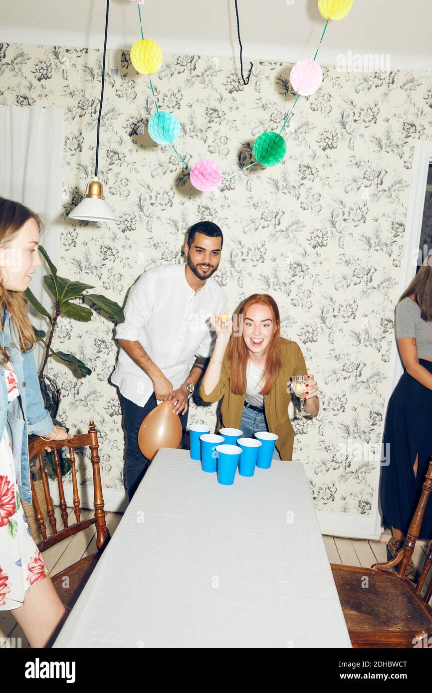 Confident young woman playing beer pong on table by male friend at