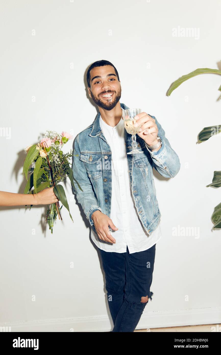 Cropped hand of woman giving flowers to smiling young man holding with drink while standing against white wall at home Stock Photo