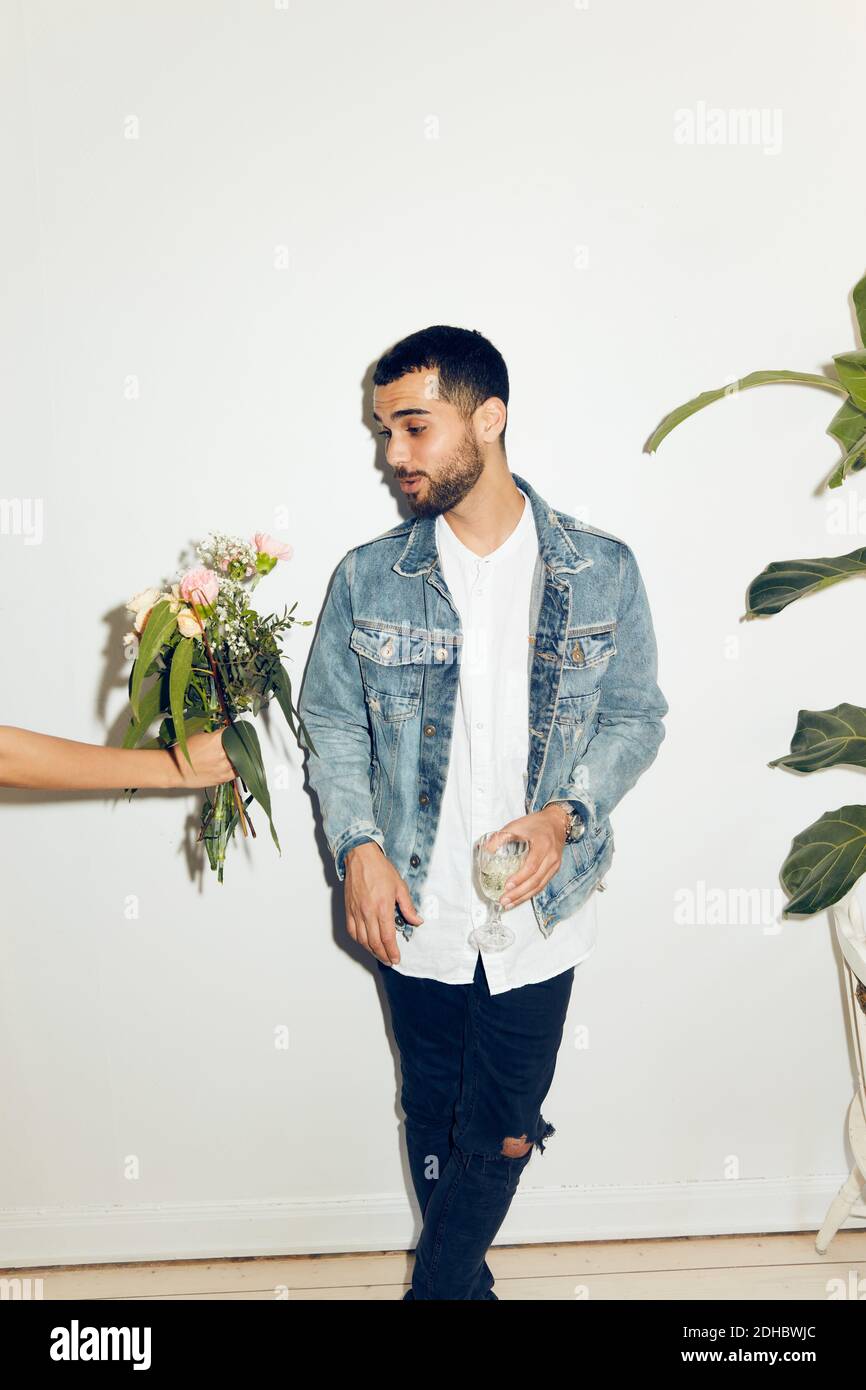 Cropped hand of woman giving flowers to young man standing with drink against white wall at home Stock Photo