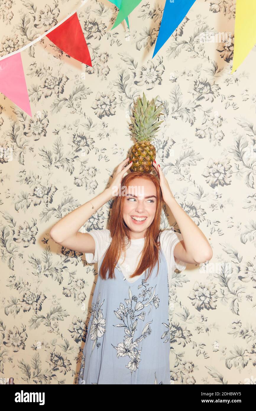 Smiling young woman carrying pineapple on head against wallpaper during party at home Stock Photo