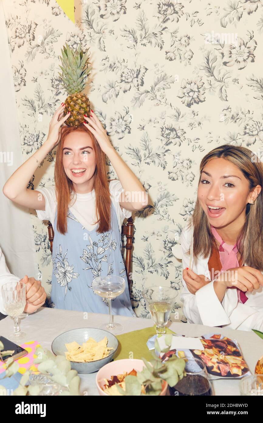 Playful young woman holding pineapple on head while sitting with friends at dining table during party in apartment Stock Photo