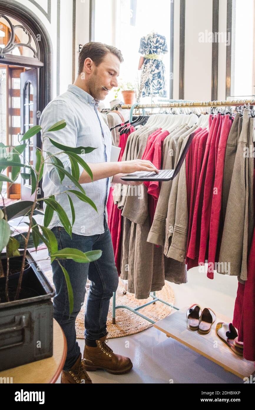 Full length of owner using laptop while standing by clothes rack in ...