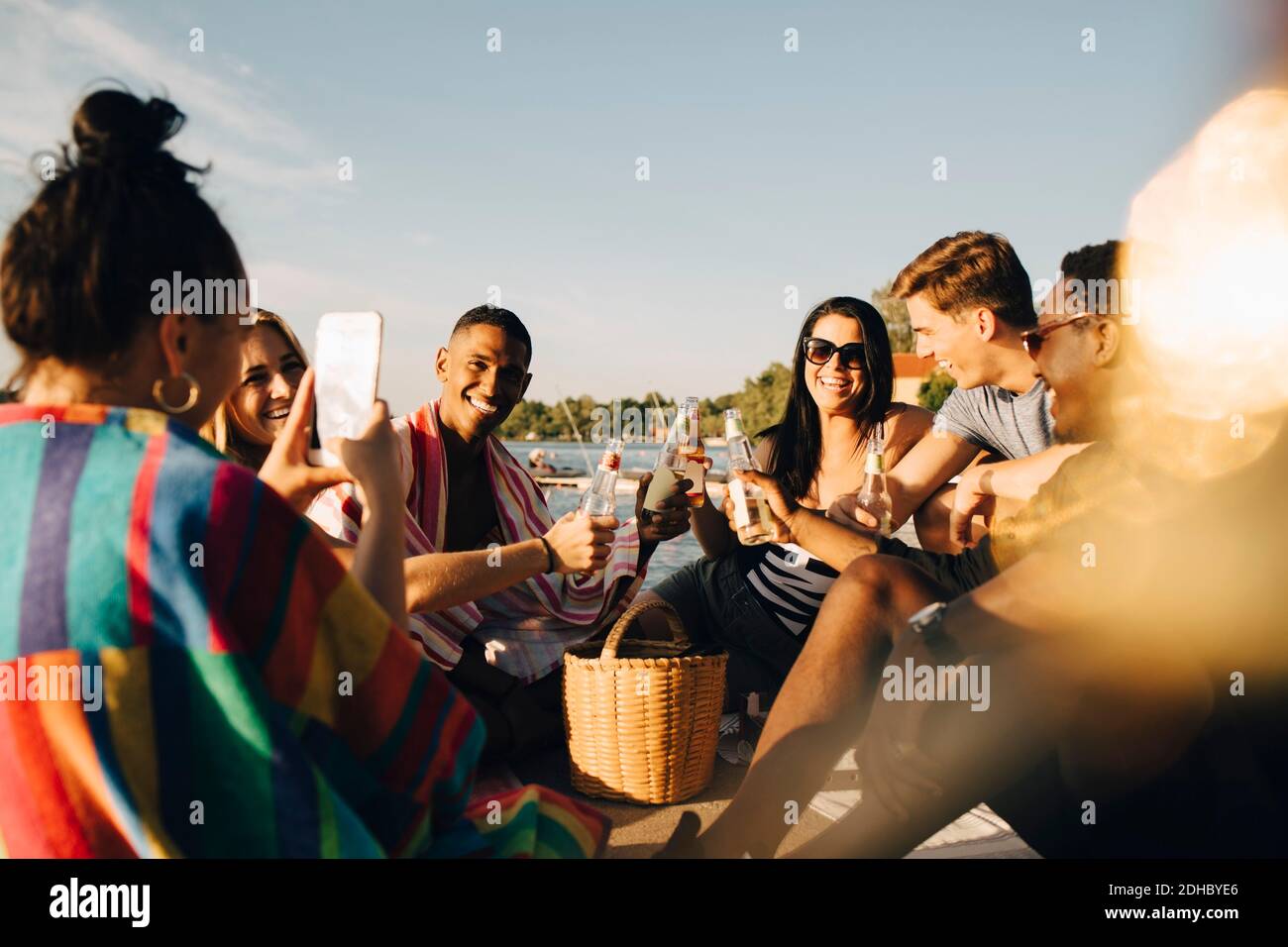 Woman photographing cheerful friends toasting drinks at jetty in summer Stock Photo