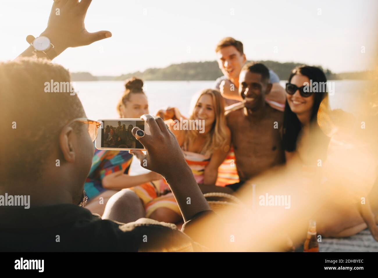 Man photographing cheerful friends on mobile phone at jetty during summer Stock Photo