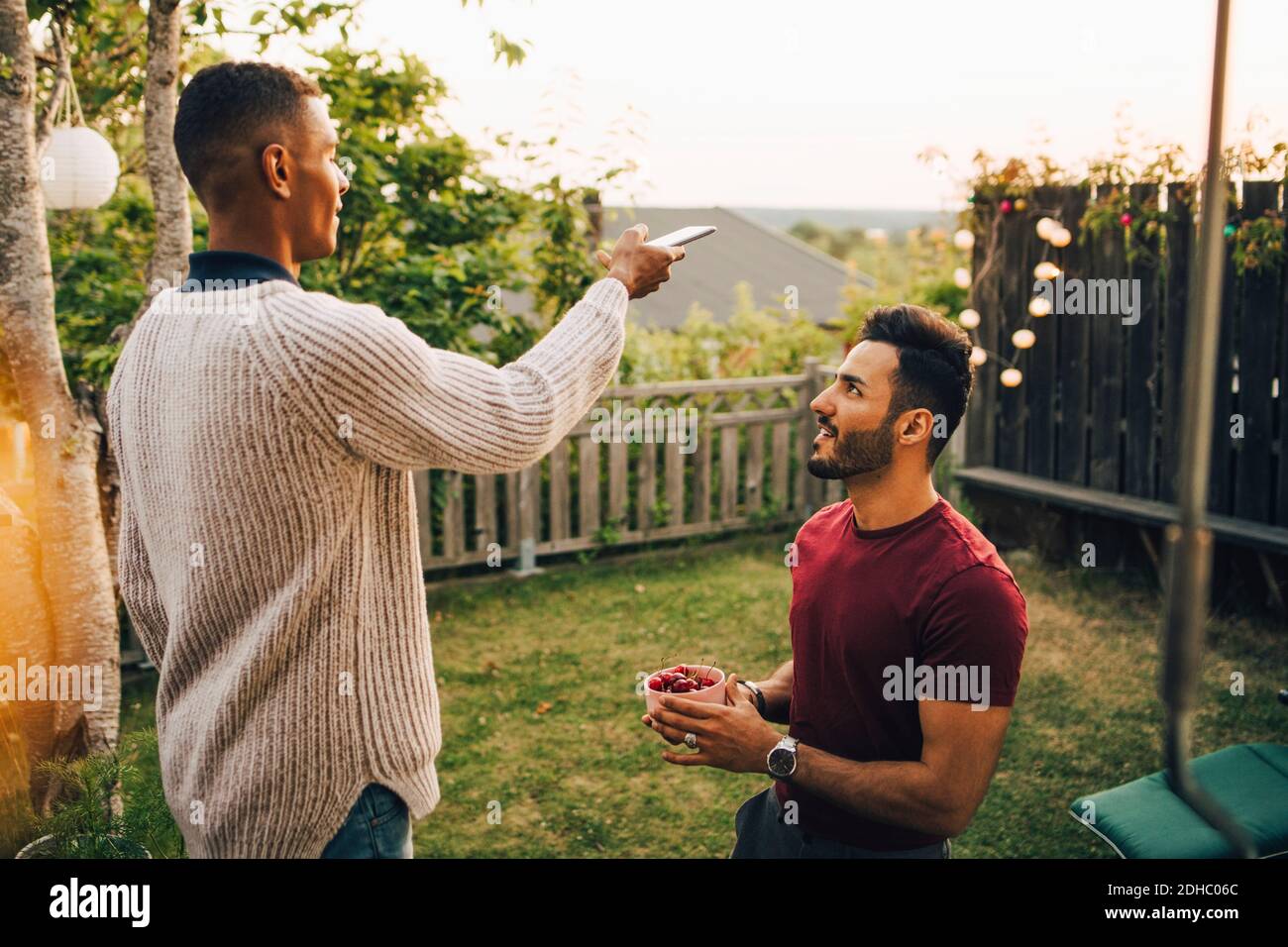 Young man photographing cherries being held by male friend in yard during sunset Stock Photo