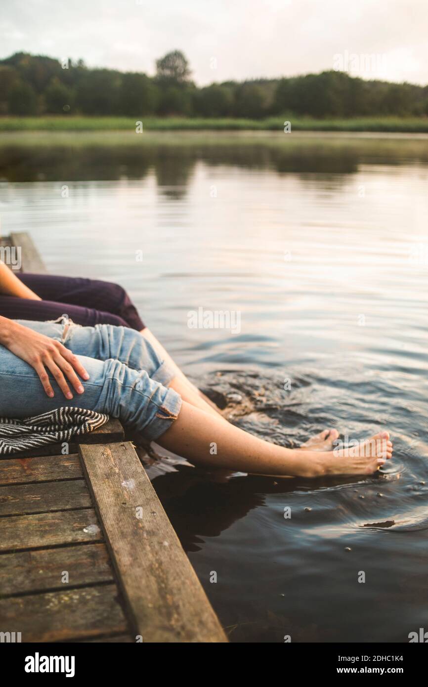 Low section of female friends dangling legs in lake while sitting on jetty Stock Photo