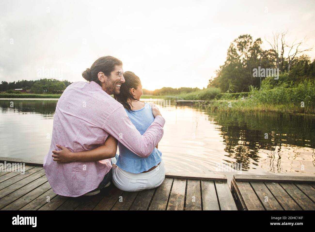 Rear view of smiling couple sitting with arms around on jetty over lake ...
