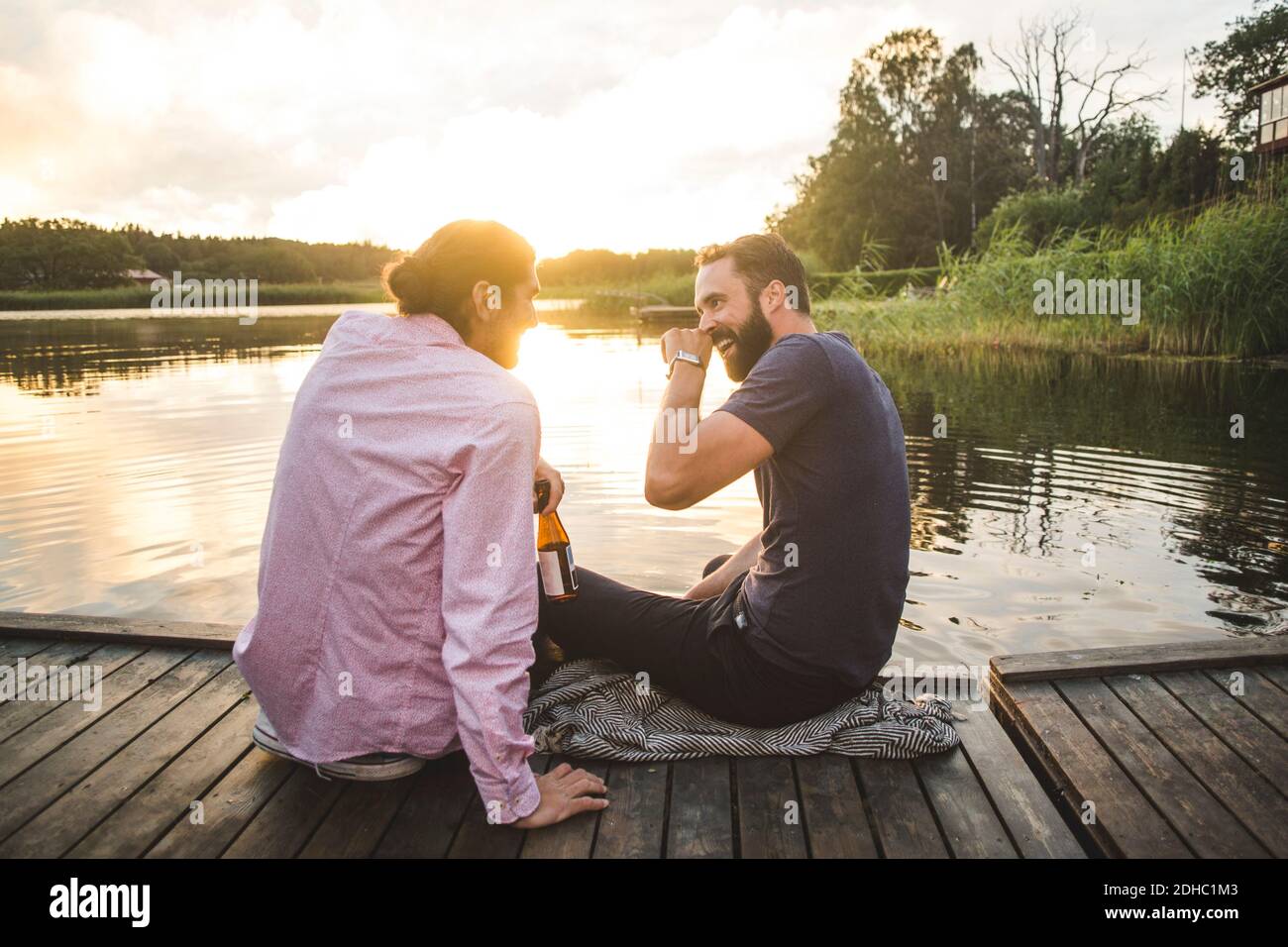 Smiling male friends talking while sitting on jetty over lake during sunset Stock Photo