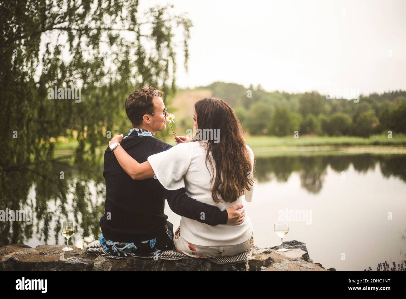 Rear view of romantic couple sitting on retaining wall against lake ...
