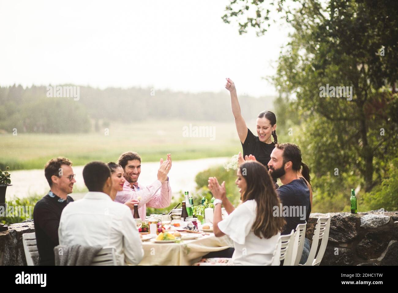 Mid adult woman standing with arm raised while friends applauding at table during dinner party in backyard Stock Photo