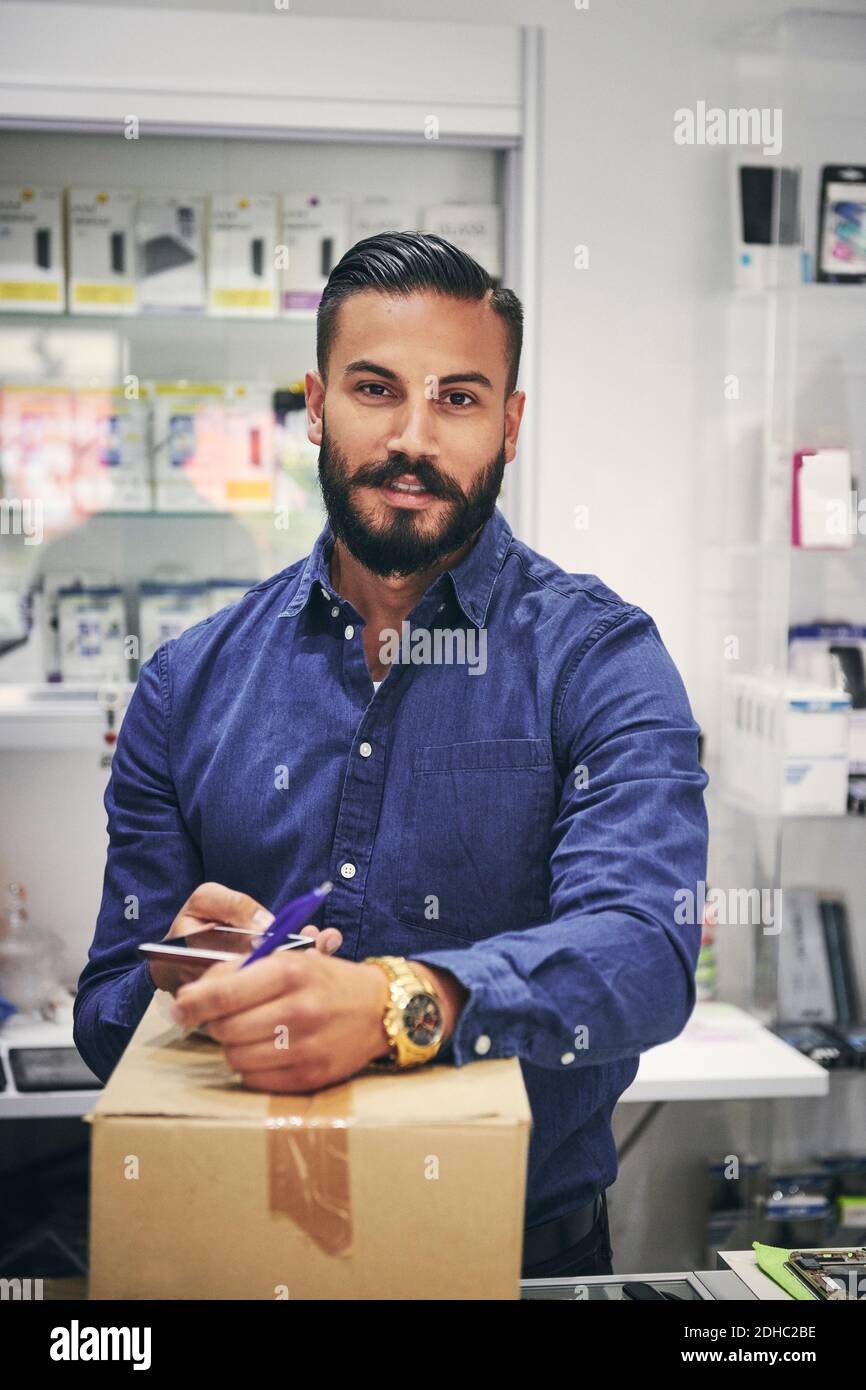 Portrait of salesman writing on box while holding mobile phone in electronics store Stock Photo