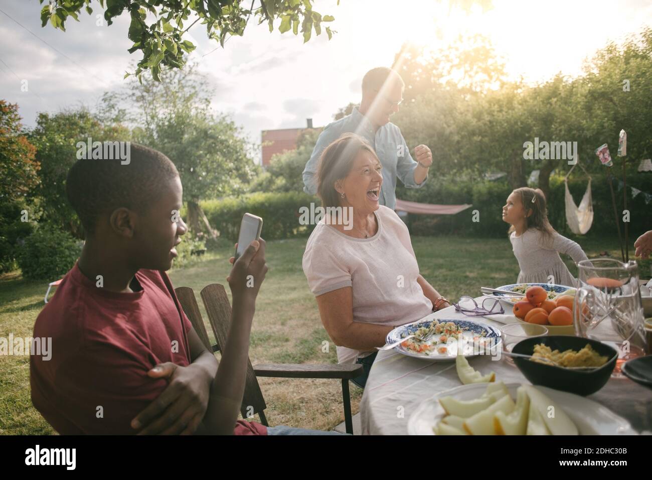 Boy photographing cheerful family enjoying lunch at table during garden party Stock Photo