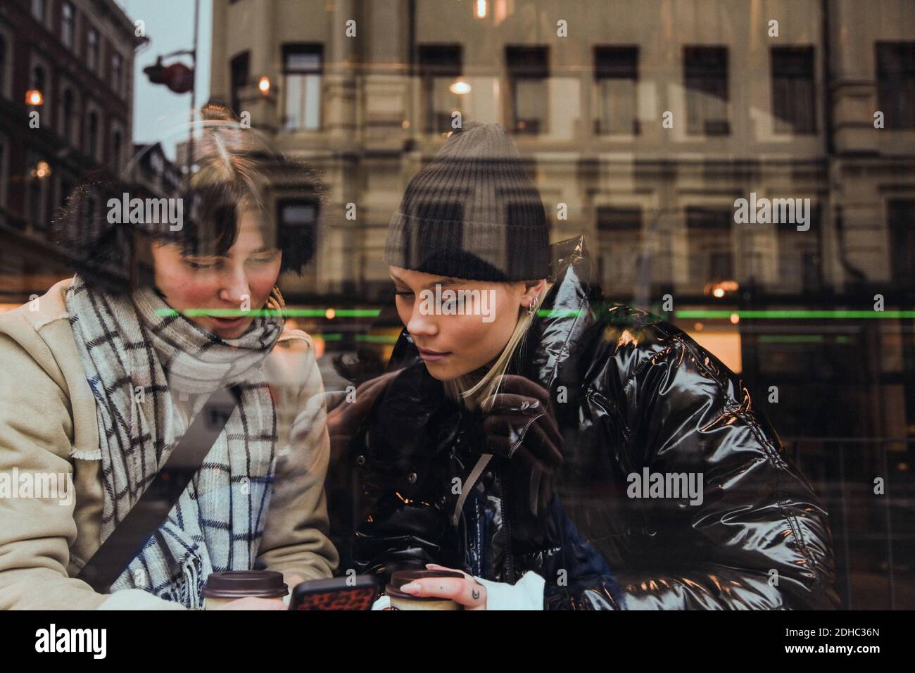 Young woman wearing warm clothing while looking at female friend using mobile phone in cafe seen through glass window Stock Photo