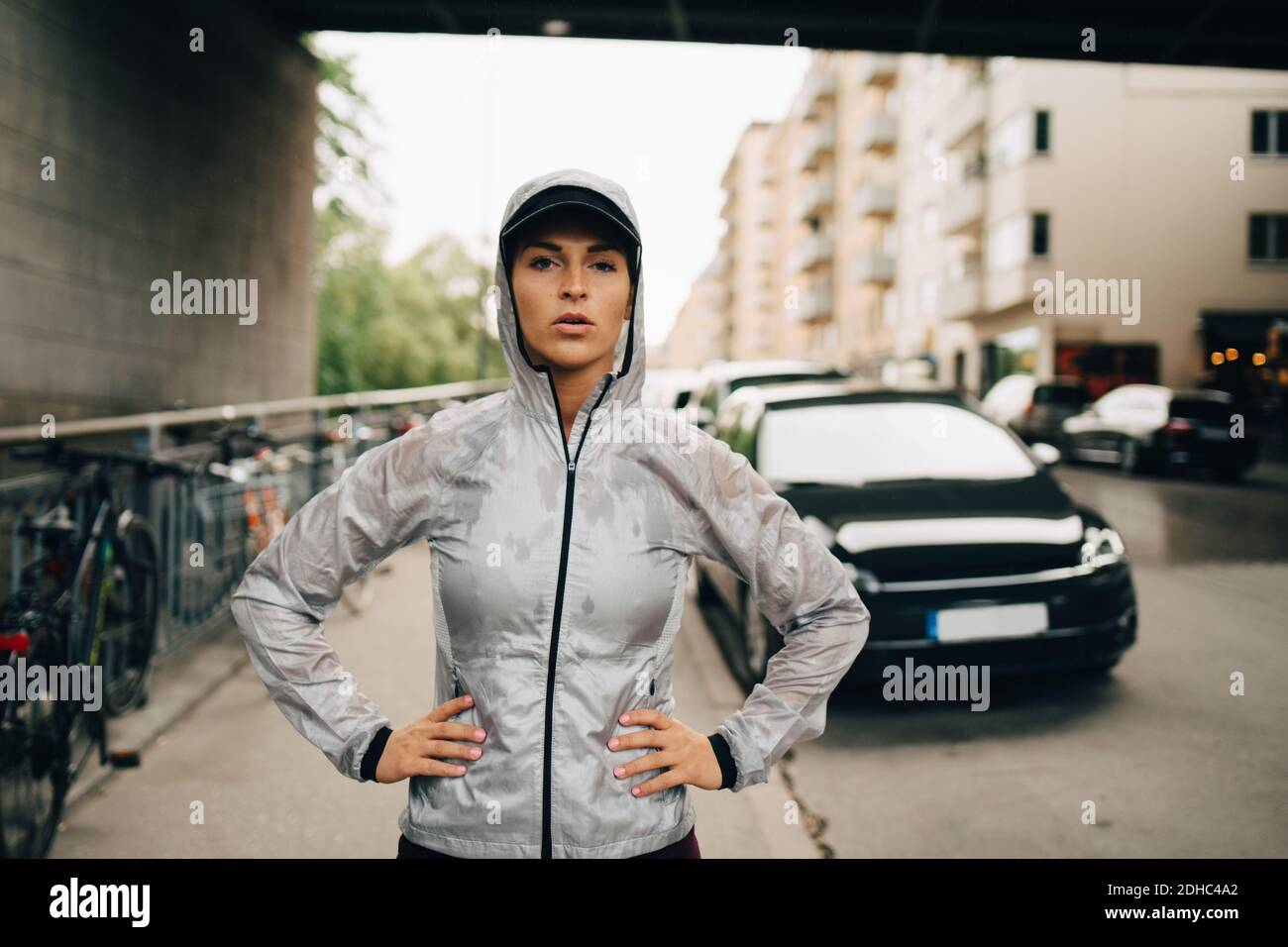 Portrait of confident female athlete in wet hooded jacket standing on sidewalk in city Stock Photo