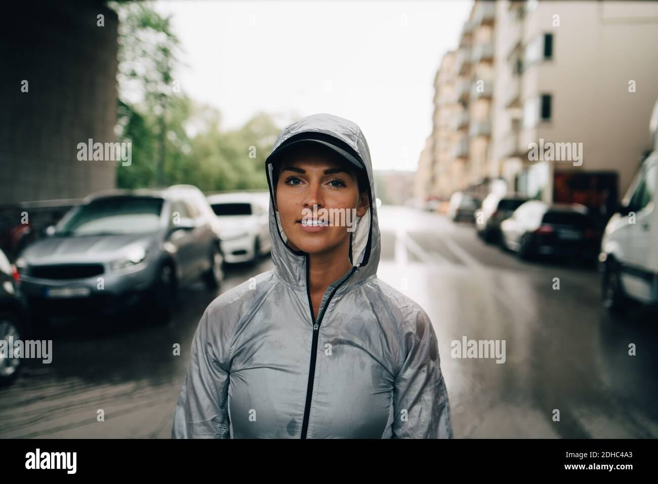 Portrait of confident female athlete in wet hooded jacket standing on street in city Stock Photo