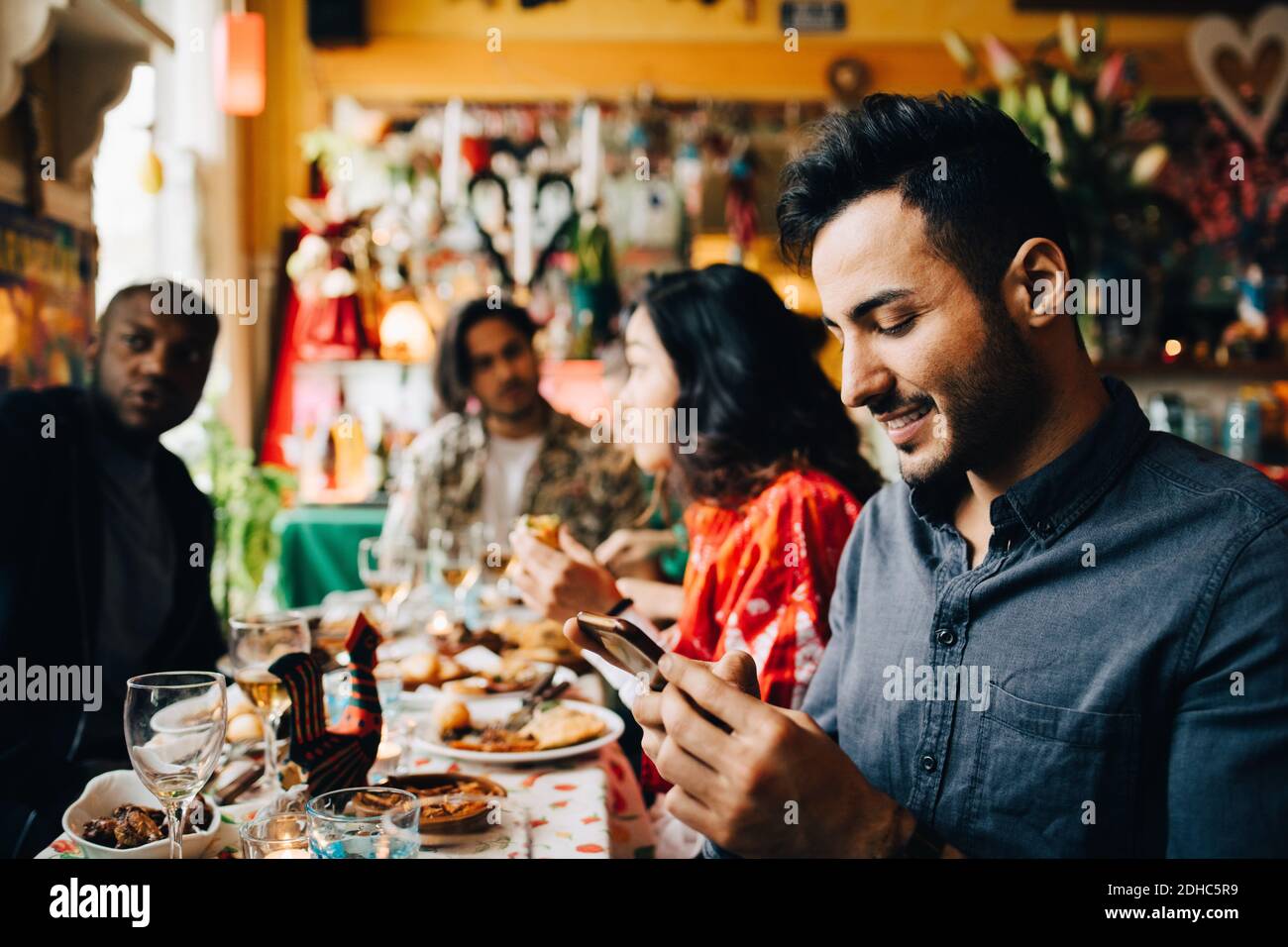 Smiling young man text messaging on smart phone while sitting with friends at table in restaurant during dinner party Stock Photo