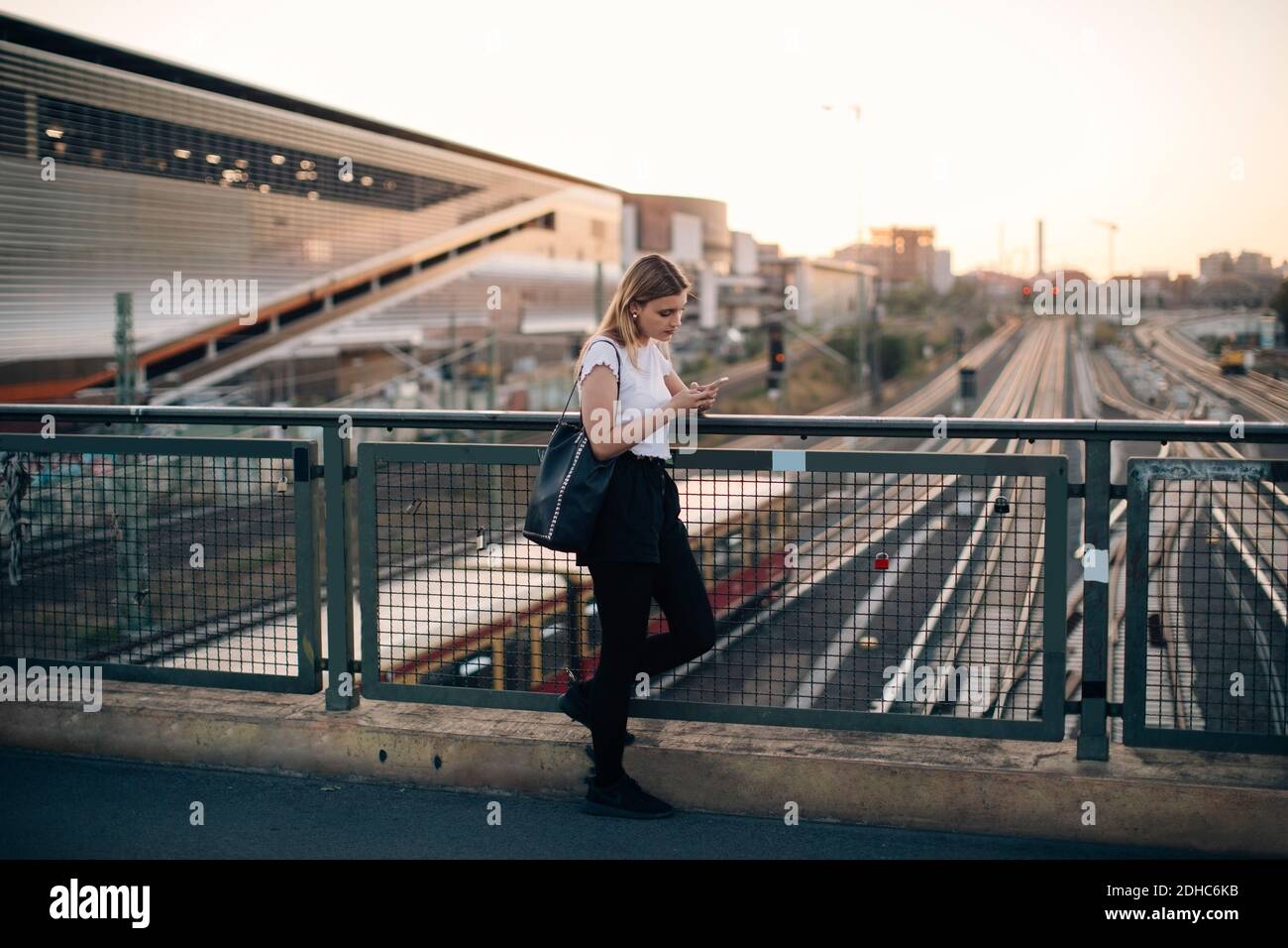 Young woman using mobile phone while leaning on railing at bridge in city during sunset Stock Photo