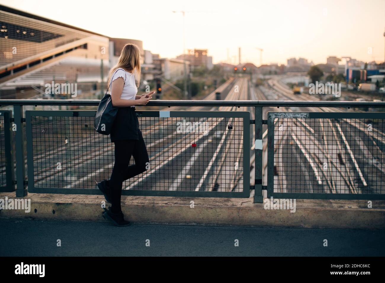 Side view of young woman standing on bridge over railroad tracks in city during sunset Stock Photo