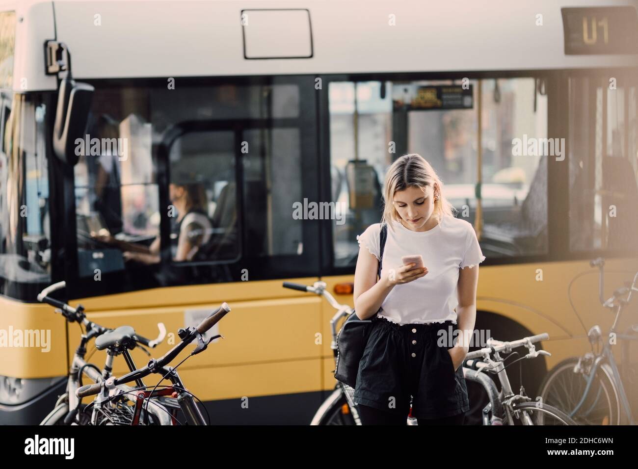 Young woman using smart phone while standing by bicycles against bus in city Stock Photo