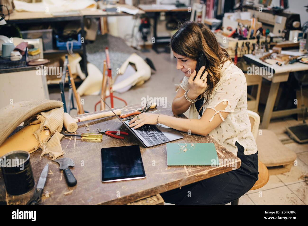 Female upholstery worker talking on phone while using laptop at workbench in workshop Stock Photo