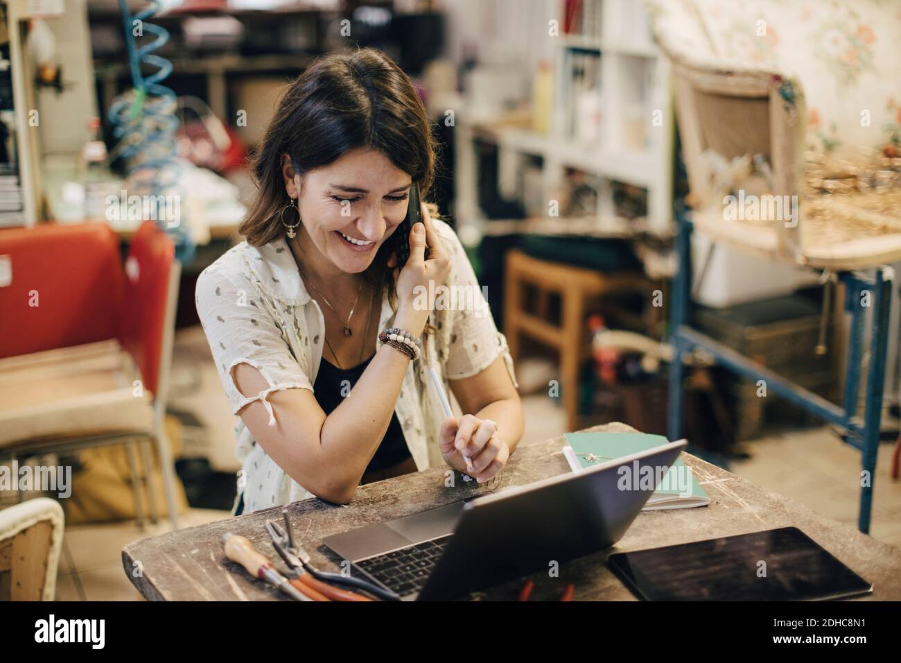Smiling female upholstery worker using laptop while talking on phone at workbench in workshop Stock Photo