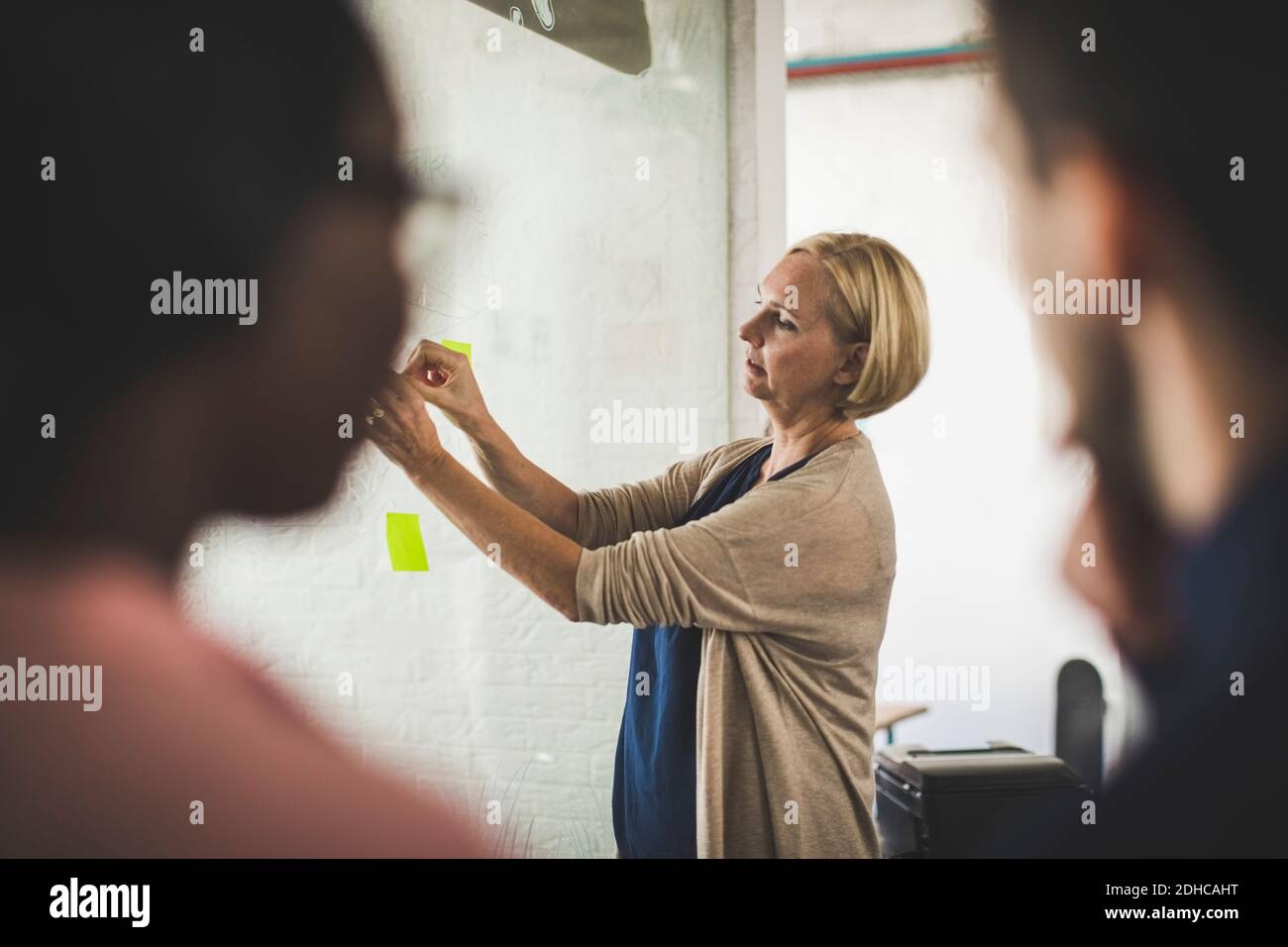 Entrepreneurs looking at female colleague sticking adhesive note on glass in creative office Stock Photo