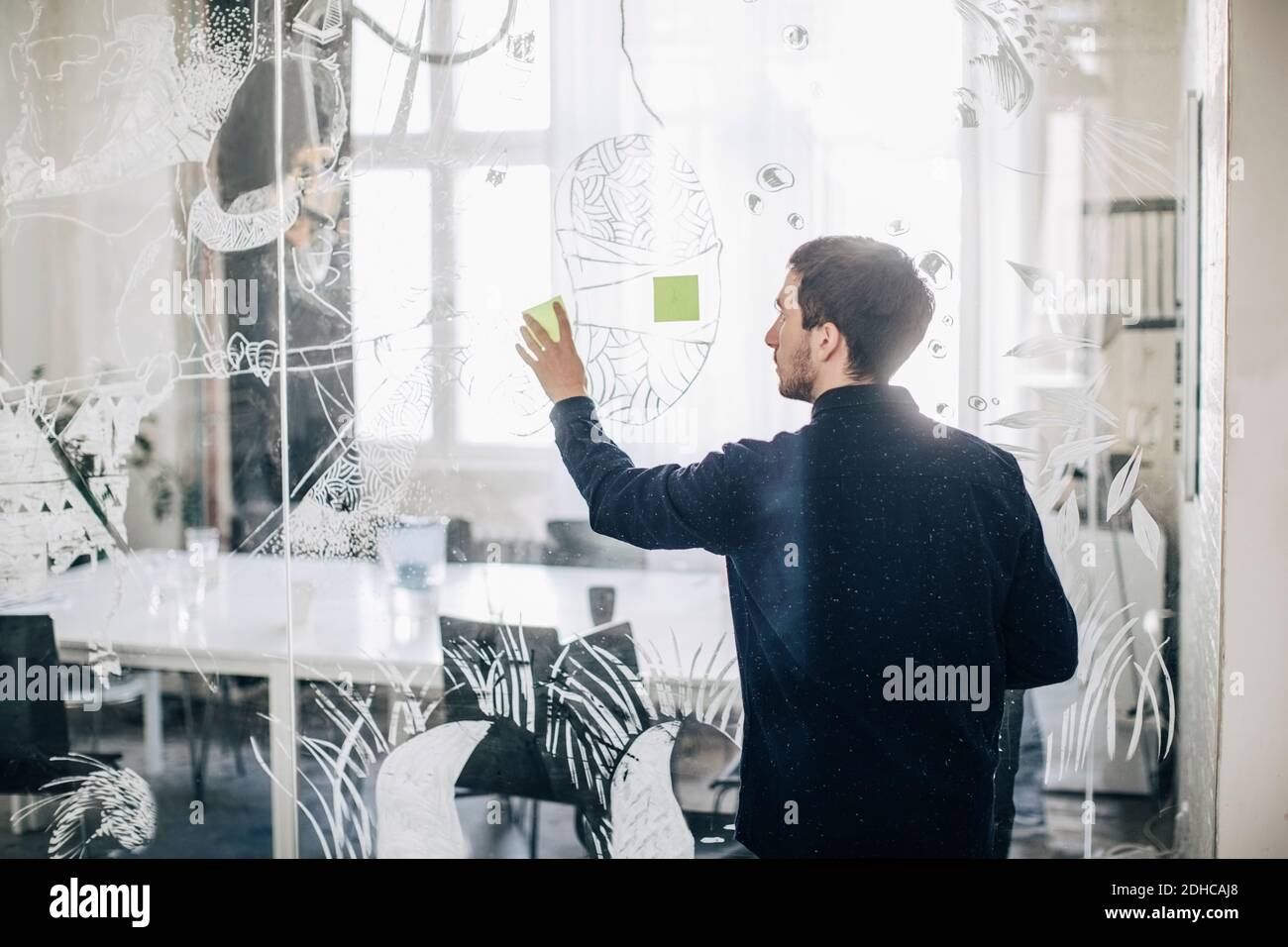 Rear view of creative businessman sticking adhesive note on glass in office Stock Photo