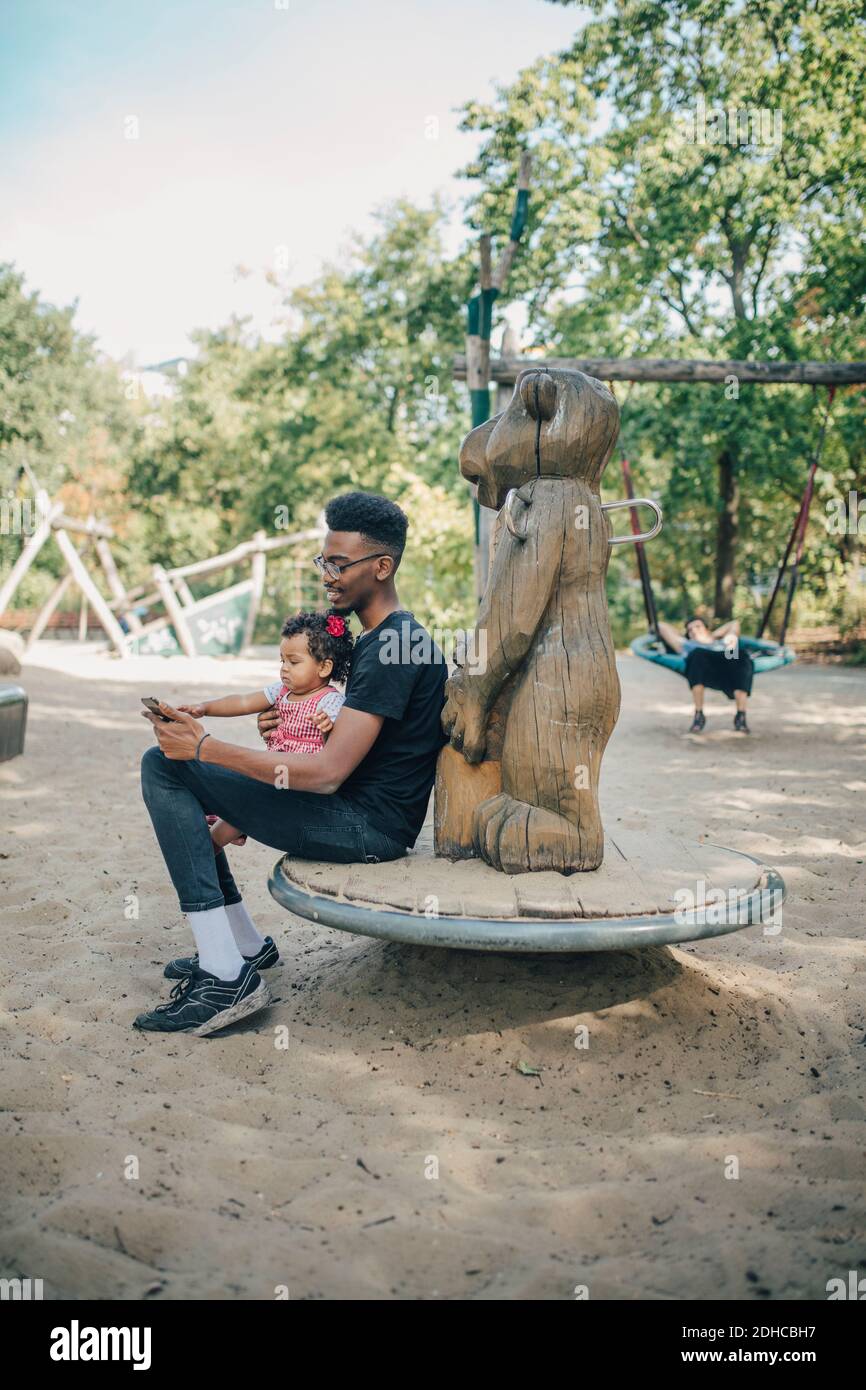 Young man using mobile phone while sitting with daughter on outdoor play equipment Stock Photo