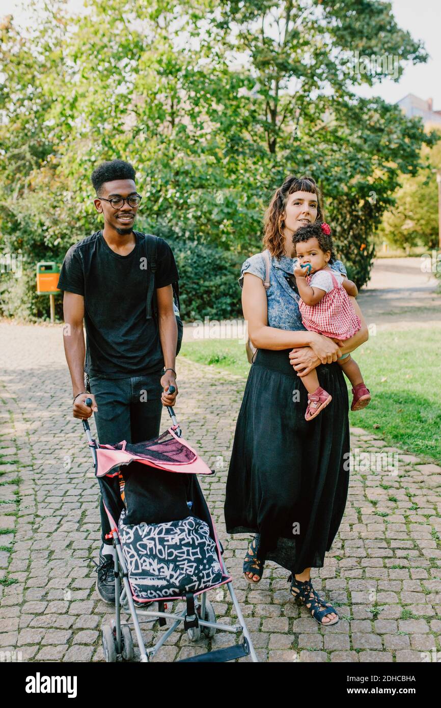Woman carrying daughter while man pushing baby stroller at footpath in park Stock Photo