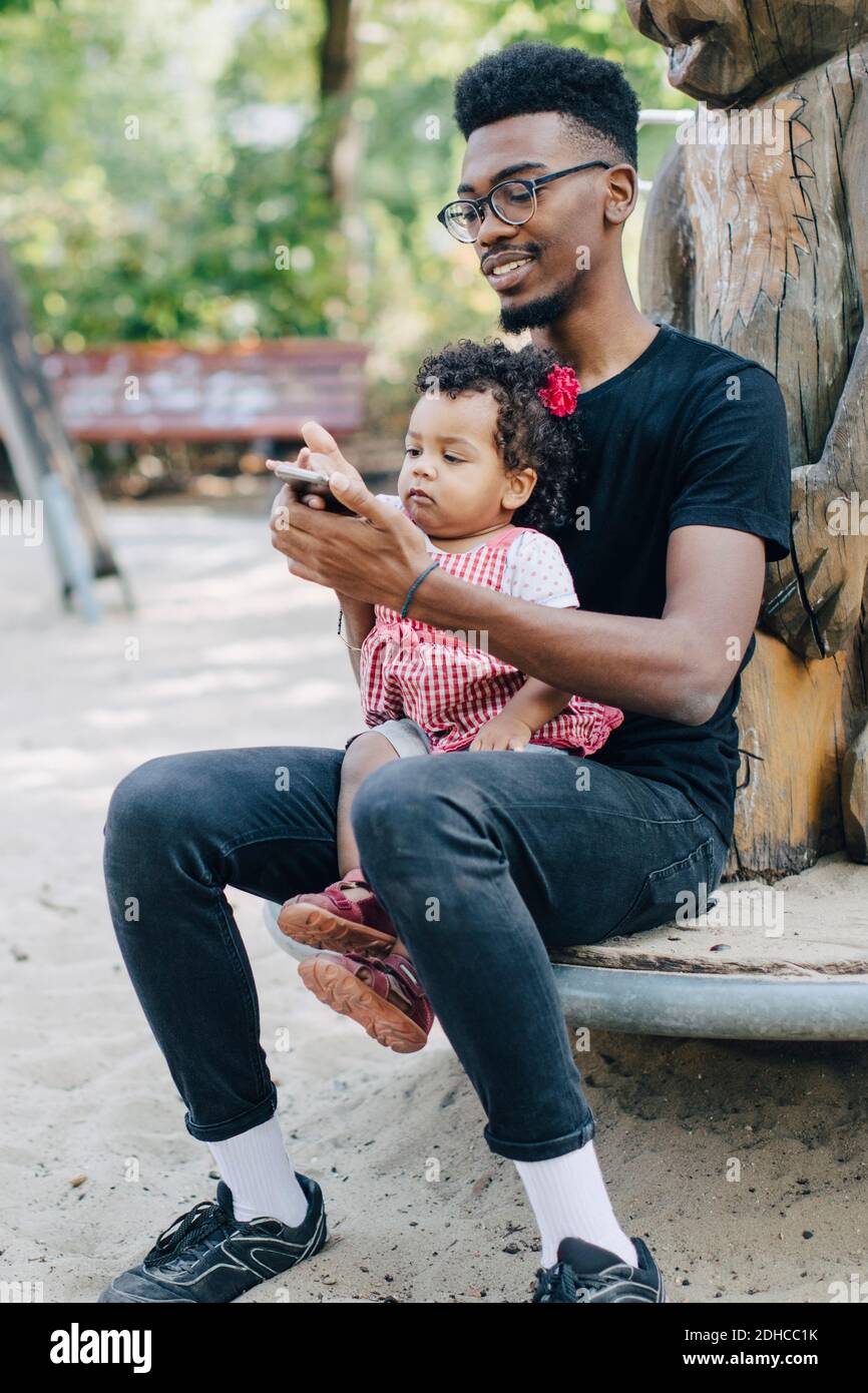 Baby girl using mobile phone while sitting with father on outdoor play equipment at playground Stock Photo