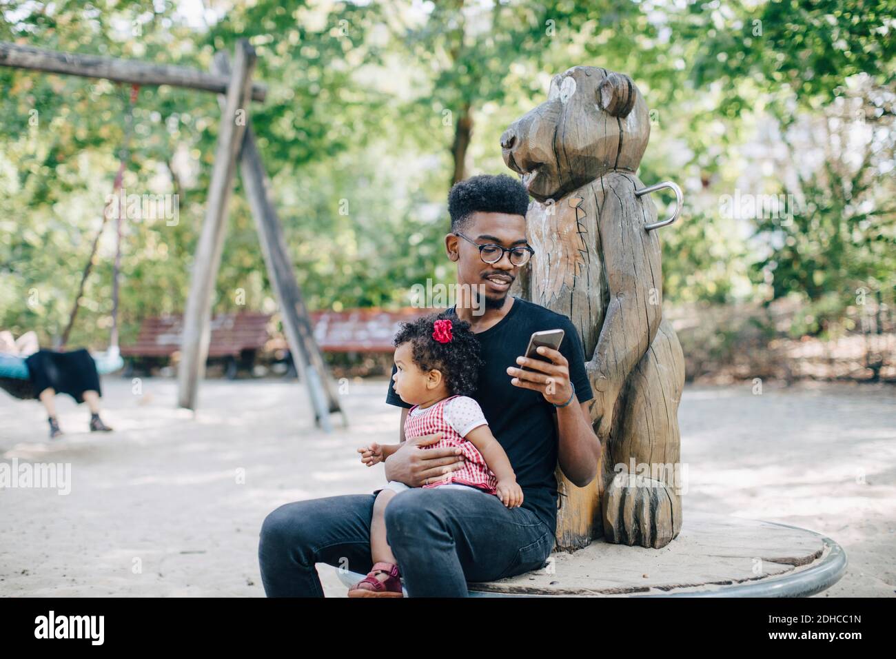 Father using mobile phone while sitting with daughter on outdoor play equipment Stock Photo