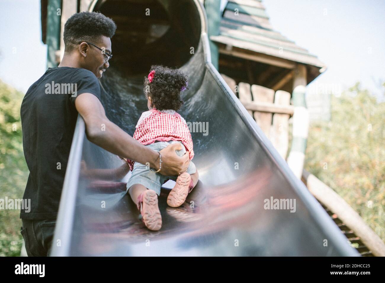 Father supporting daughter crawling up on slide at playground Stock Photo