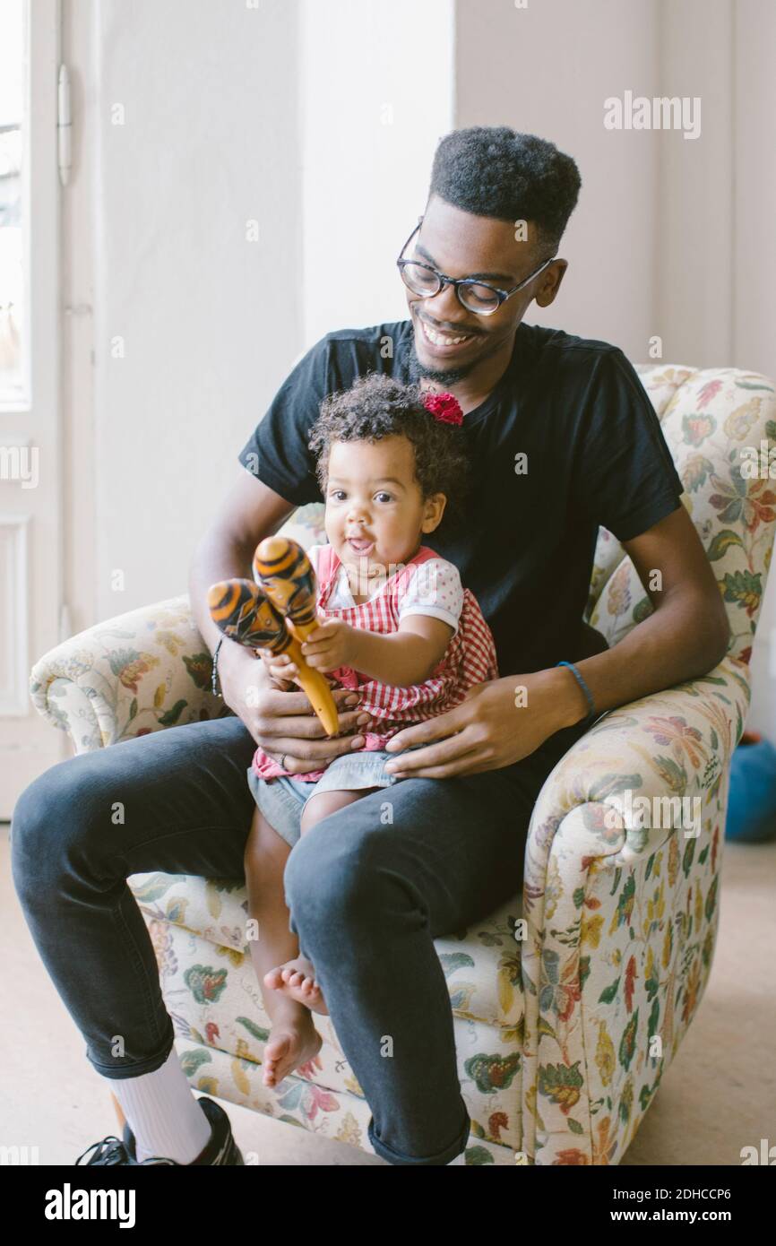 Playful baby girl playing holding maracas while sitting with father on armchair at home Stock Photo