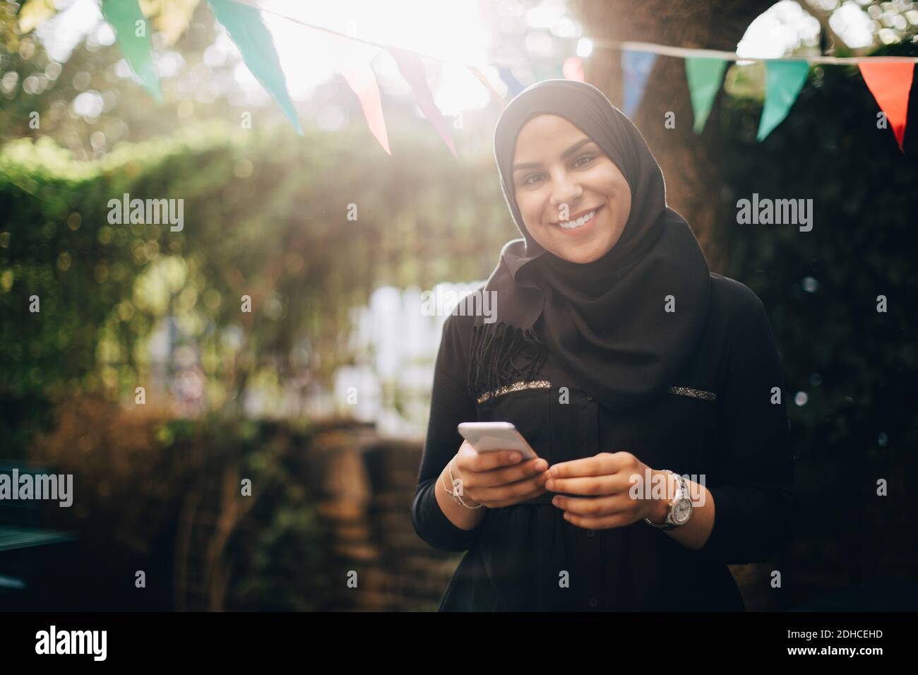 Portrait of smiling woman in hijab holding smart phone at backyard ...