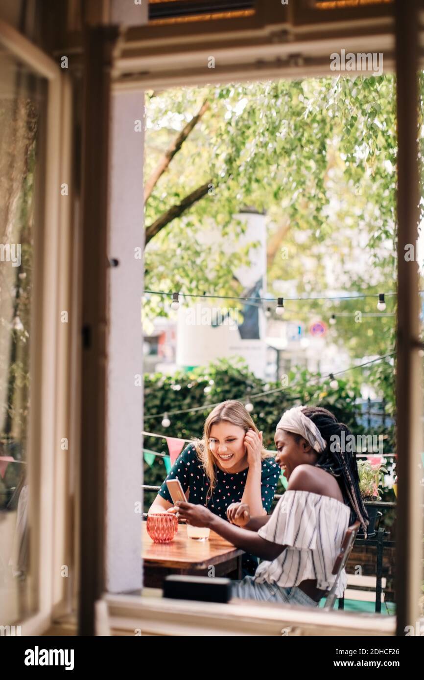 Happy multi-ethnic female friends looking at mobile phone seen through window Stock Photo