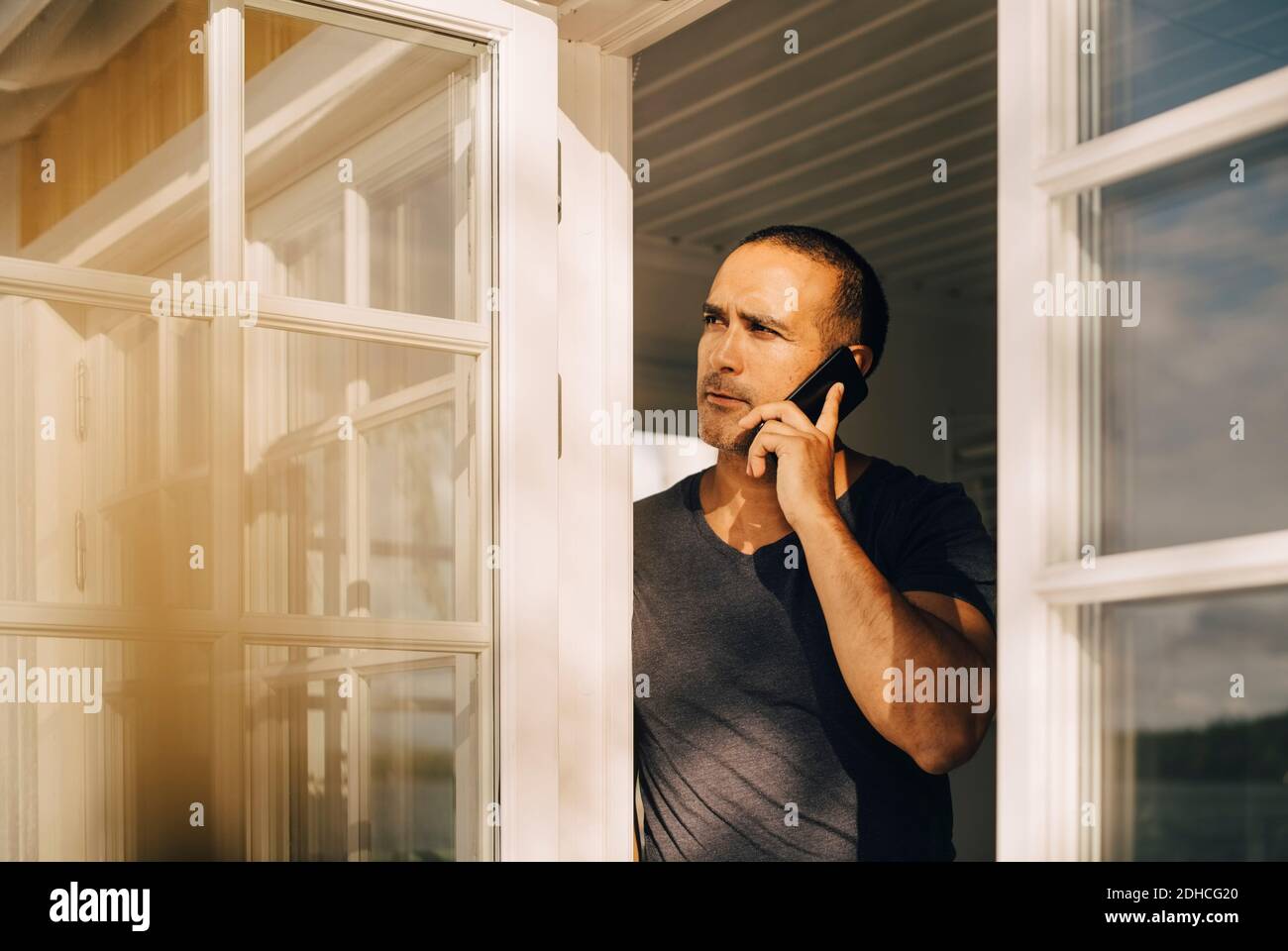 Thoughtful man talking on smart phone while standing at window Stock Photo