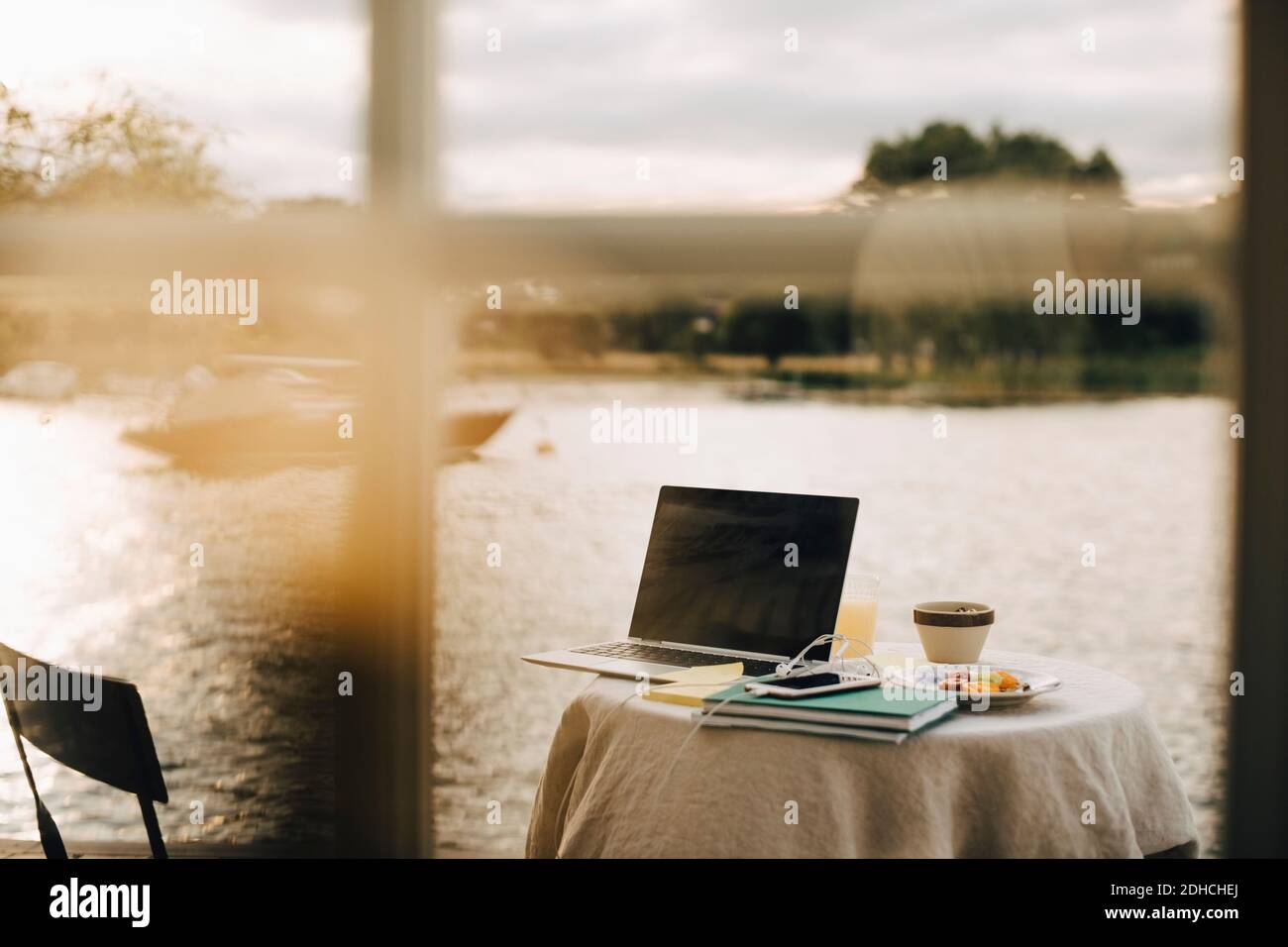 Laptop and smart phone with books by breakfast on table against lake seen through window at holiday villa Stock Photo
