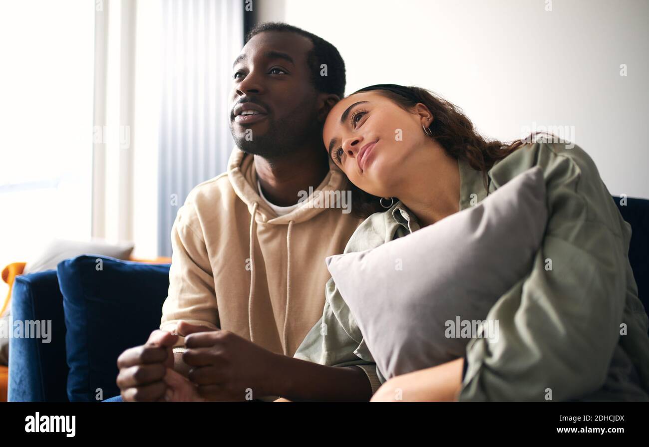 Loving young mixed ethnicity couple cuddling whilst sitting on sofa at home watching or streaming romantic movie on tv together Stock Photo