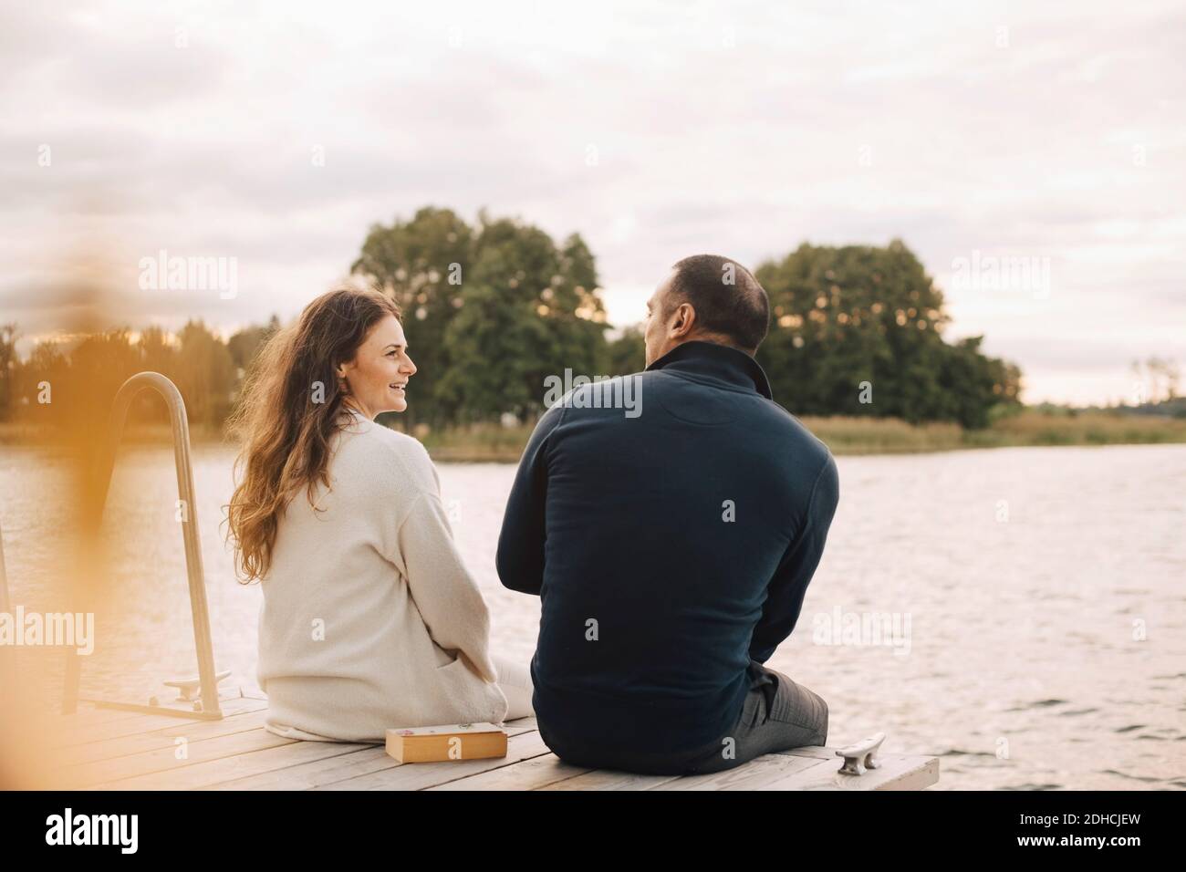 Smiling couple talking while sitting on jetty by lake against sky Stock Photo