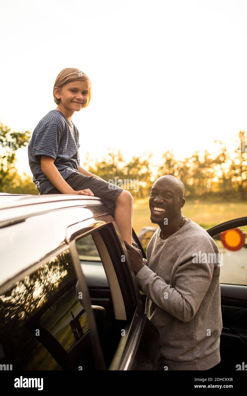 Portrait of smiling man by daughter sitting on car roof during sunset at park Stock Photo