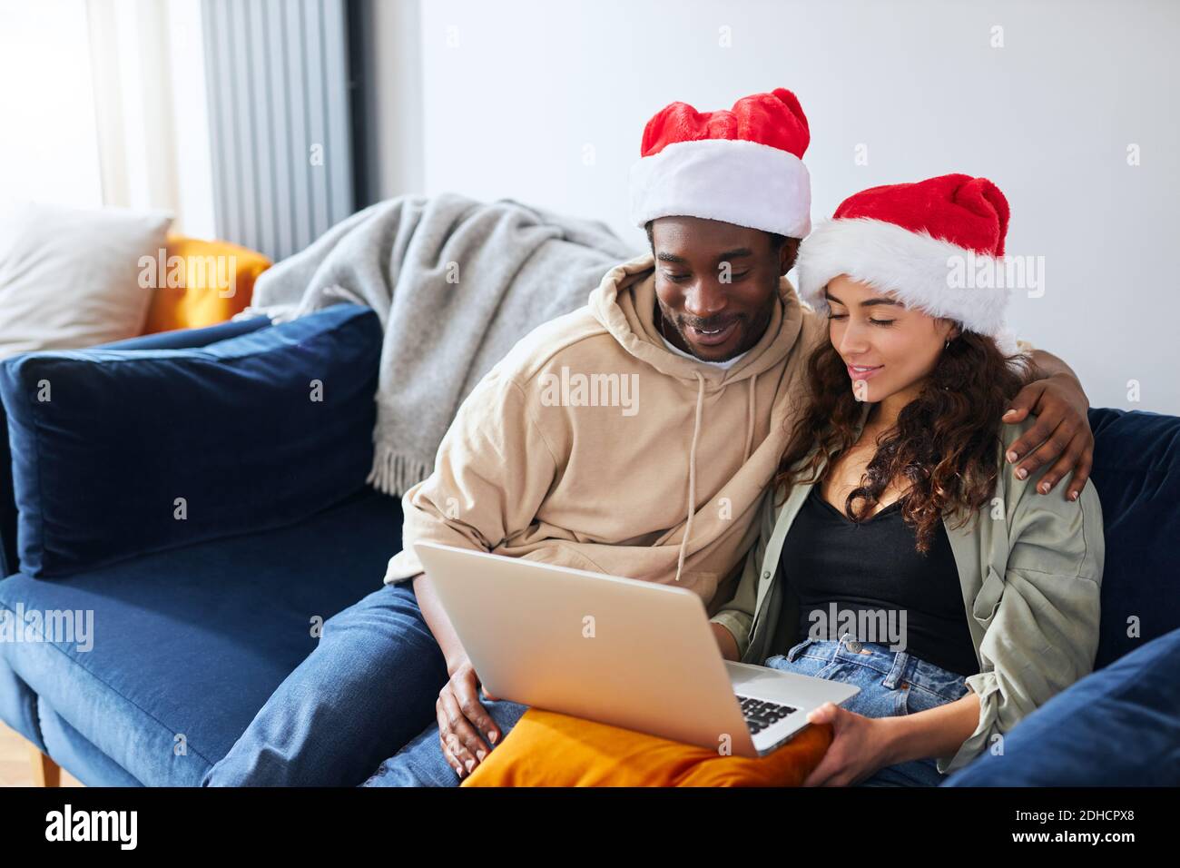 Young mixed ethnicity couple wearing santa hats sit side by side on sofa at home shopping for christmas presents online using laptop Stock Photo