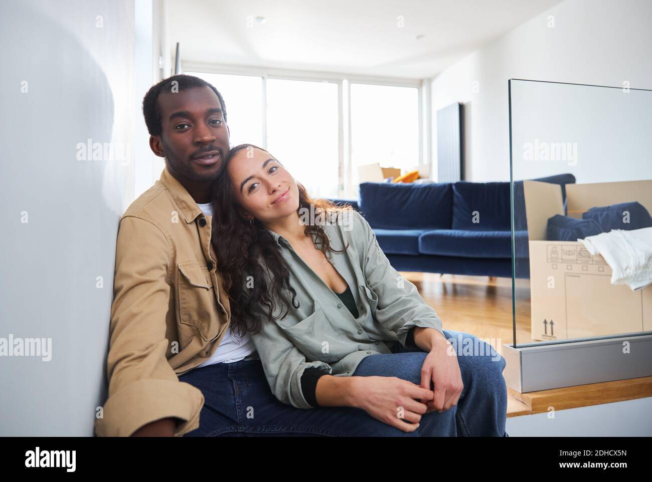 Portrait of young mixed ethnicity couple celebrating moving day in lounge of new home sitting on floor surrounded by removal boxes together Stock Photo