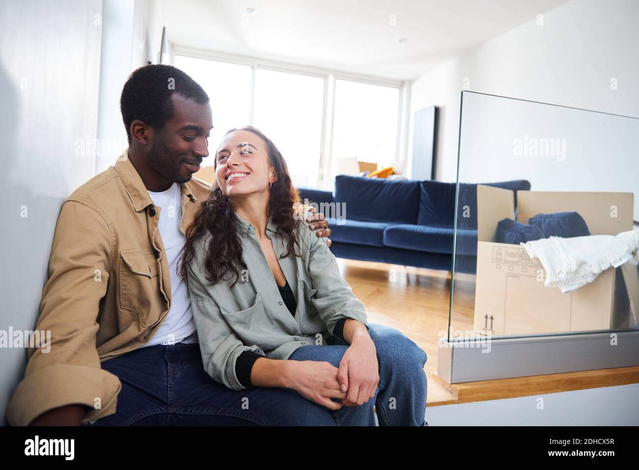 Young mixed ethnicity couple celebrating moving day in lounge of new home sitting on floor surrounded by removal boxes together Stock Photo