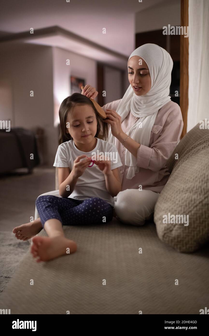Adult Muslim woman combing hair of girl while sitting on comfortable sofa at home Stock Photo