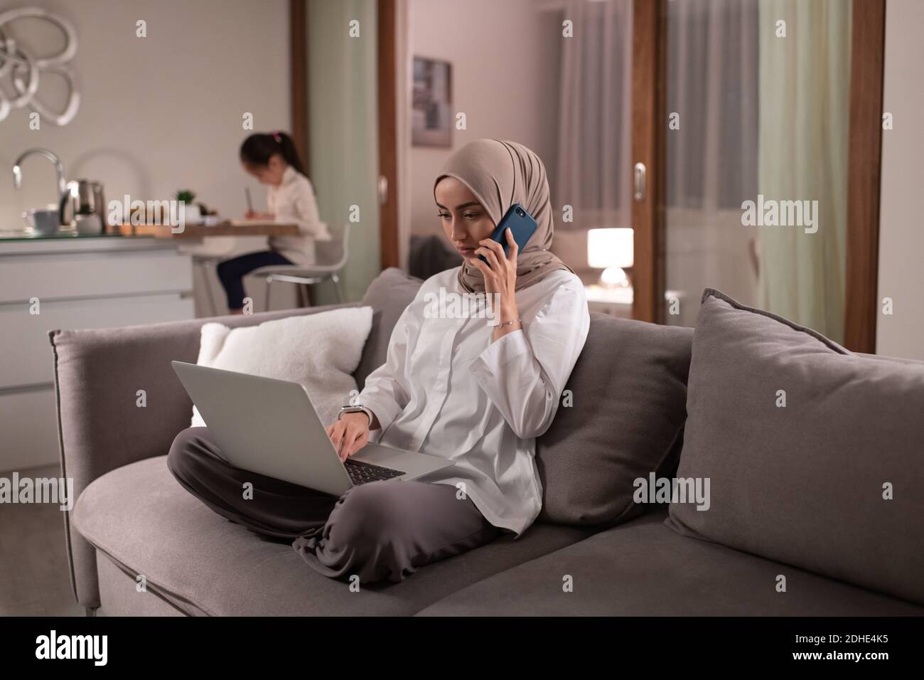 Muslim woman using laptop and speaking on phone near drawing daughter at home Stock Photo