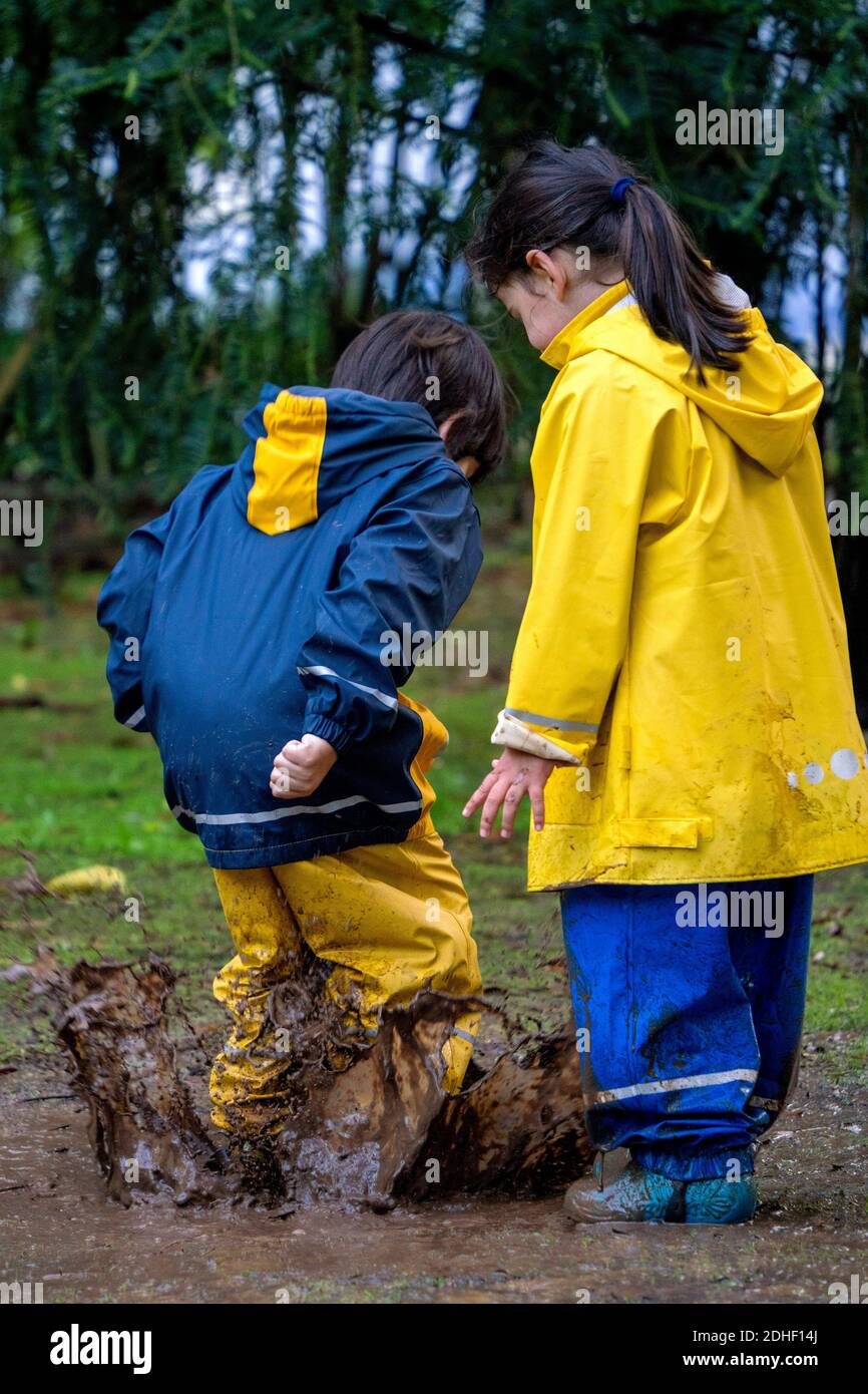 Two young kids having fun playing outside jumping in a mud puddle wearing coloful raincoats Stock Photo