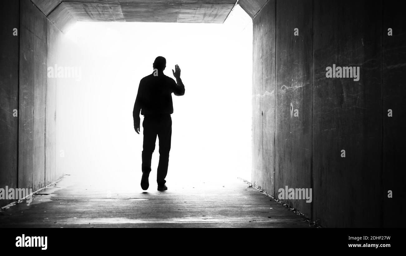 A single male in a dark tunnel exiting and walking towards a bright white light while waving goodbye or hello. Stock Photo